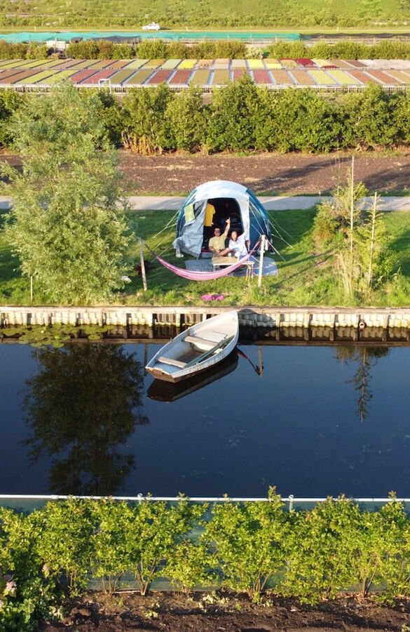 Luchtfoto van een glamping-tent tussen bomen en groen veld, met een bootje in het kanaal eronder en tulpenvelden op de achtergrond