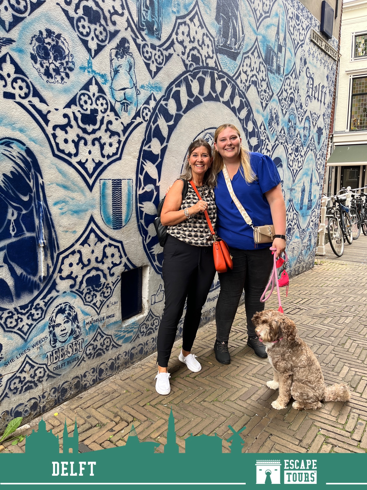 Twee vrouwen en een klein hondje poseren voor een grote muur met blauw-wit decoratief muurschildering