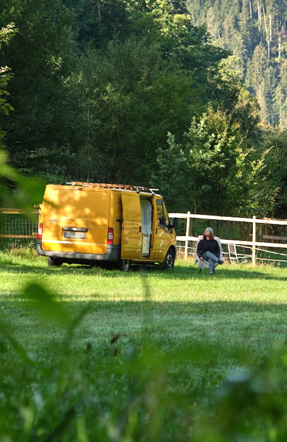 Gele campingbus geparkeerd in een bergweide met een persoon ernaast en bebost berglandschap op achtergrond