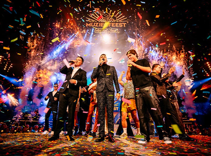 Groep performers op groot podium tussen vuurwerk en confetti tijdens muziekfestival