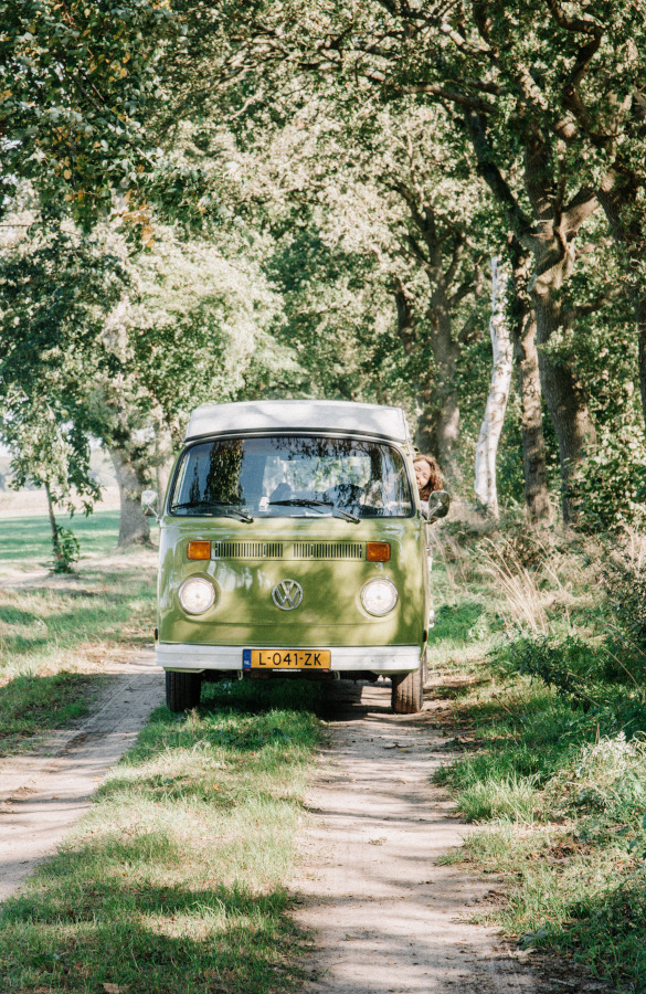 Groene vintage Volkswagen busje onder grote bomen langs een bospad met persoon erin zichtbaar