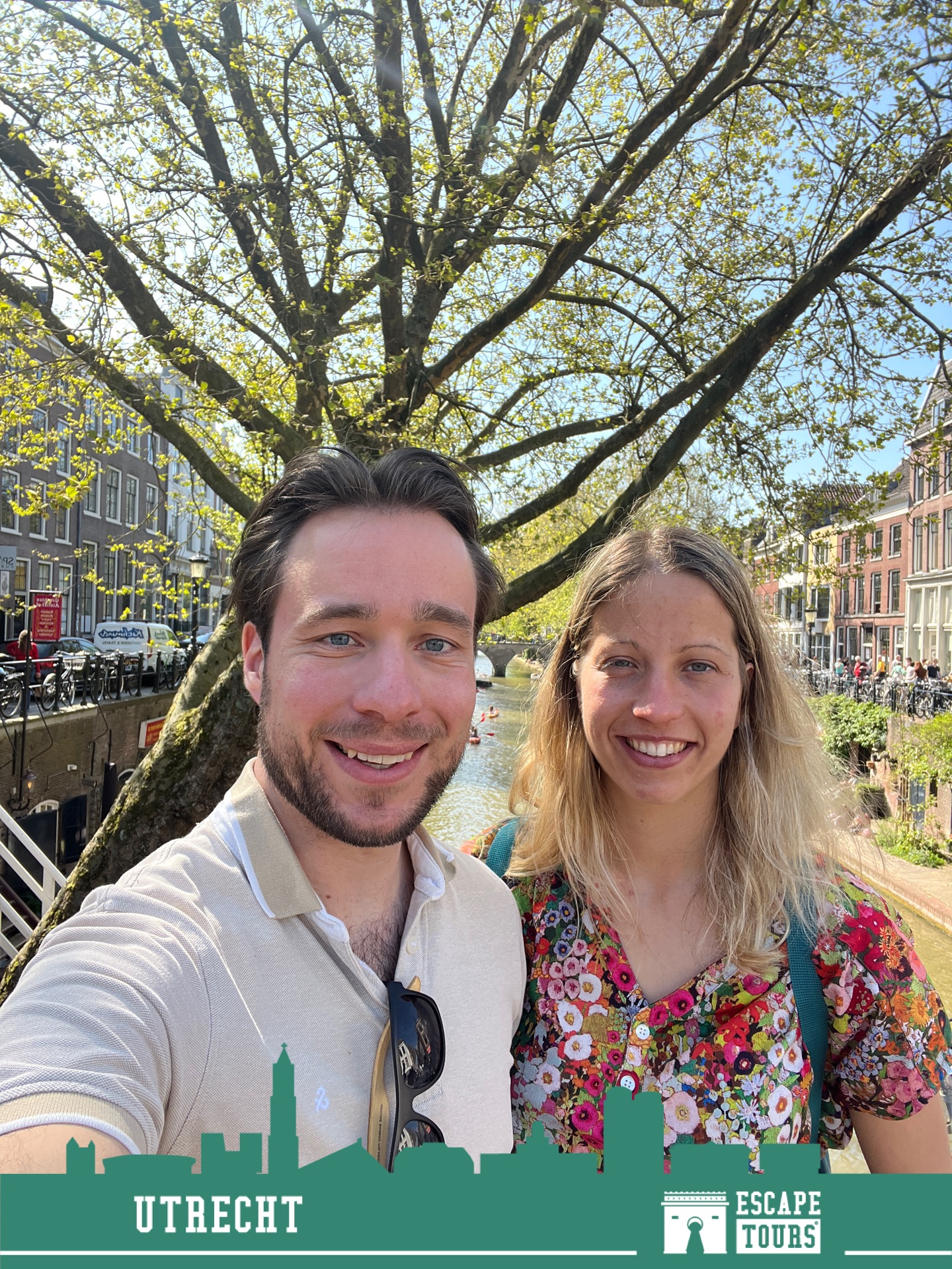 Man en vrouw poseren samen onder een grote boom langs een kanaal met fietsen en toeristen op de achtergrond